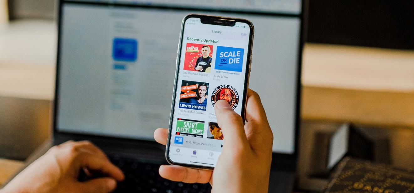 A person sitting at a computer browses popular podcasts on a phone.