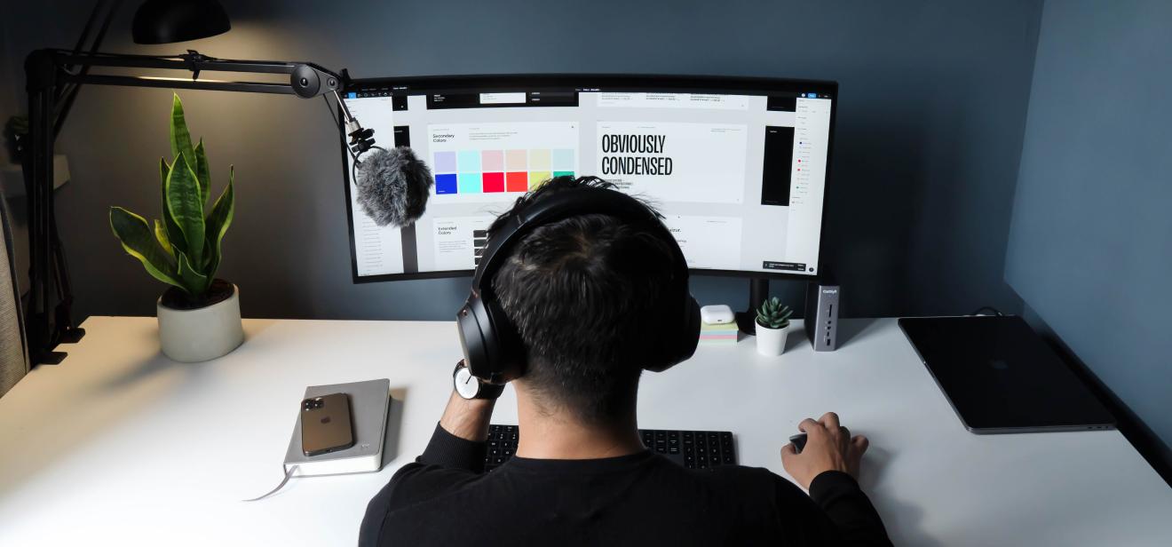 A student wearing headphones sits before a professional mic and a desktop monitor with colors schemes and graphics on the screen.