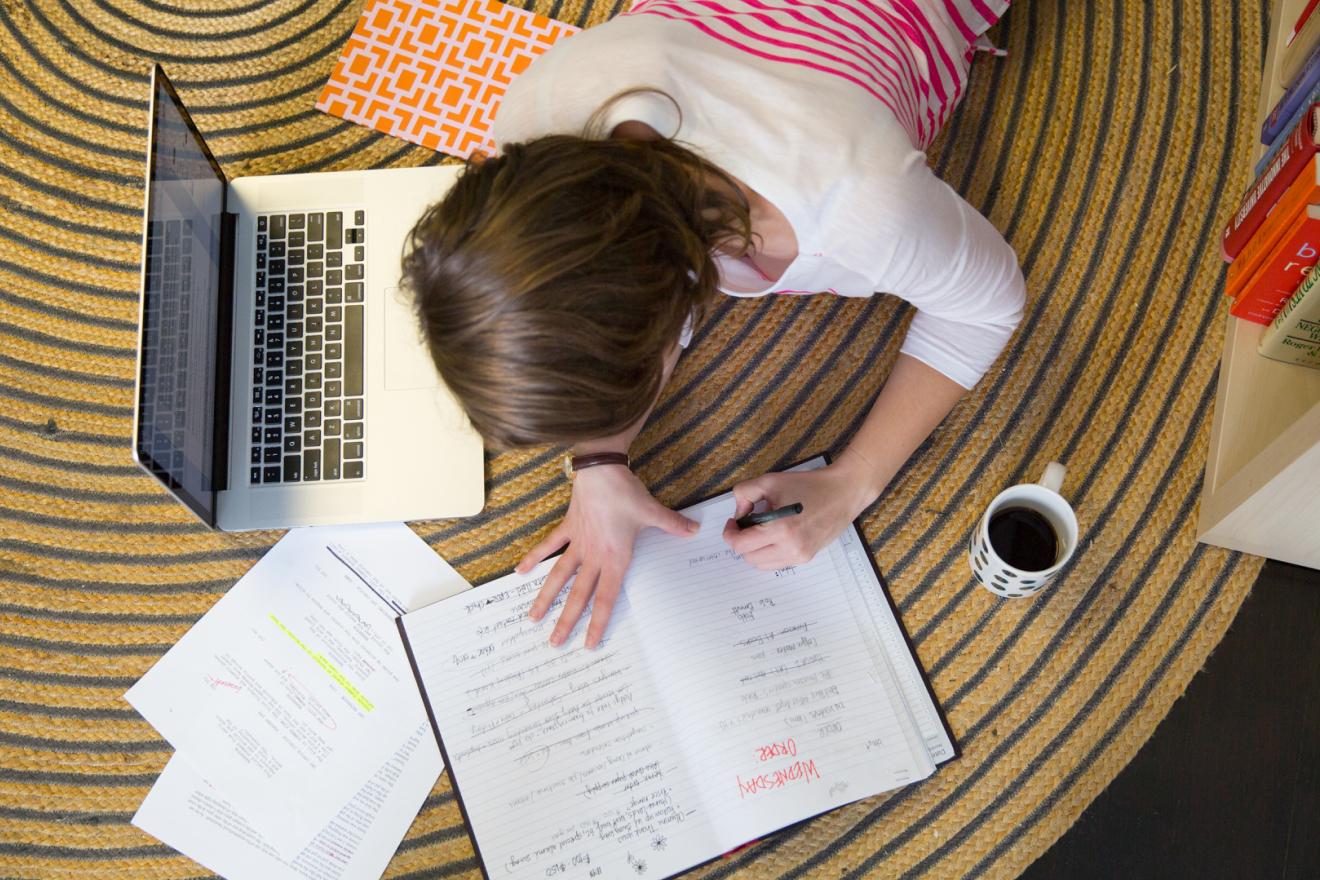 A student lays on the floor with a laptop and some coffee while writing notes in a notebook.