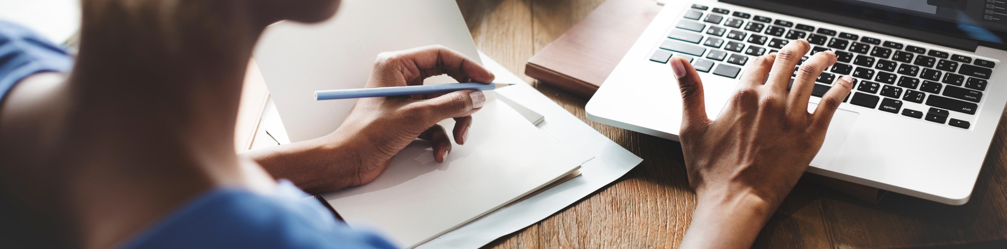 A student holds a pen above a piece of paper while using a laptop.