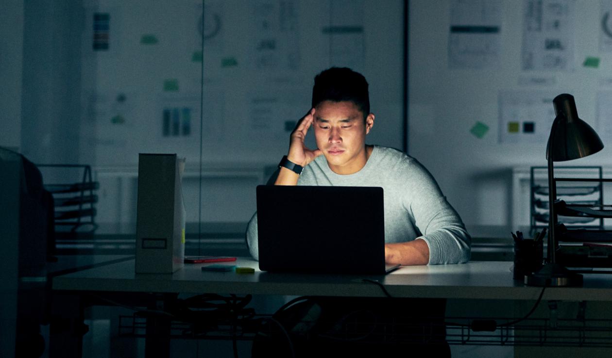 Student sits at a desk and stares intently at a laptop screen, resting his head on his hand, while the screen illuminates his face.