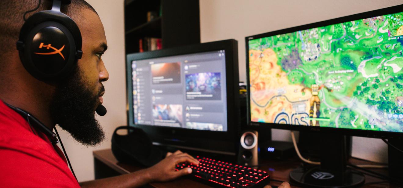 A student wearing a headset works on a PC game on two monitors with a gaming keyboard.