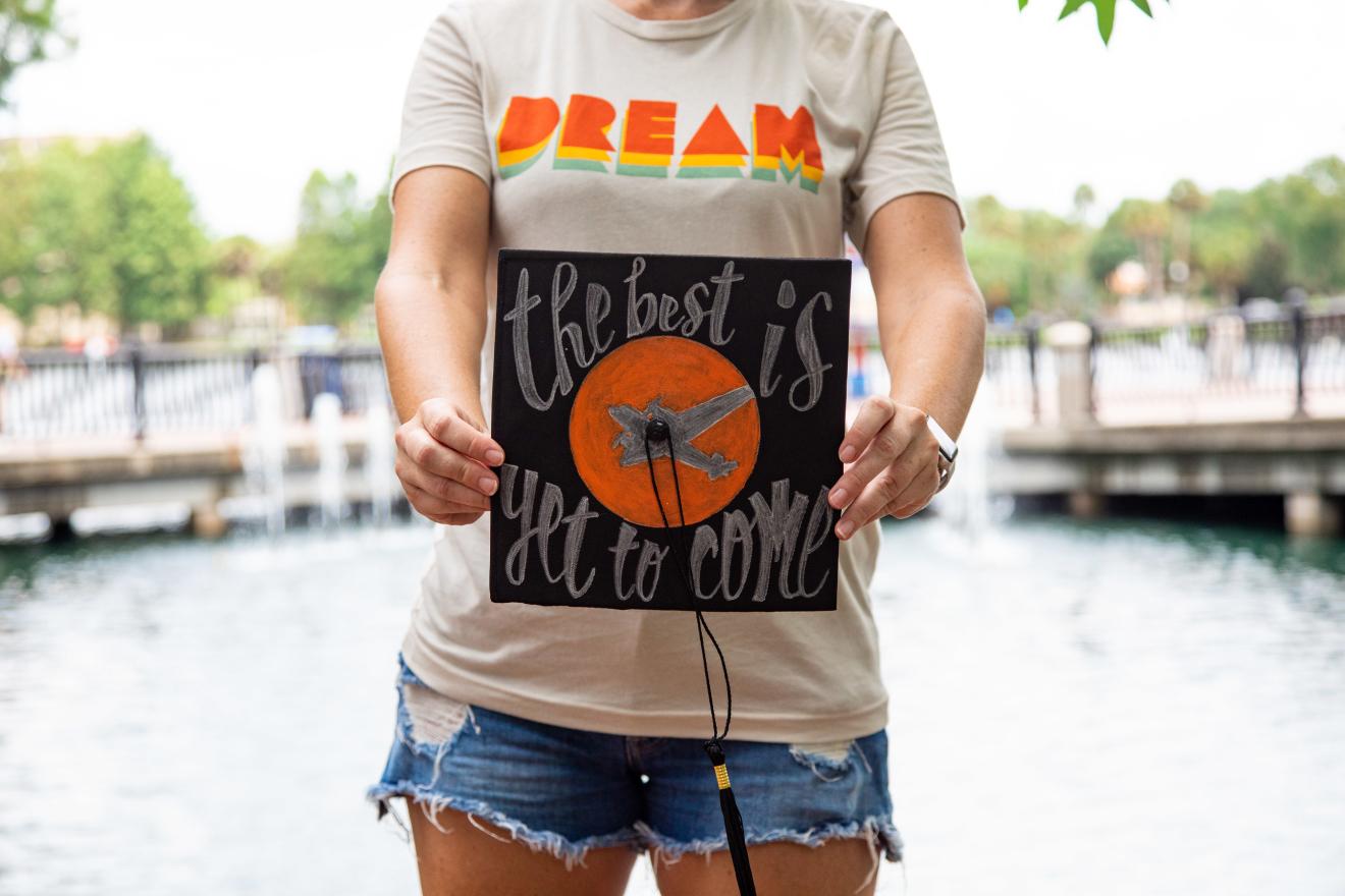 A student wearing a DREAM shirt holds a graduation cap with the Full Sail logo and the words the best is yet to come painted on the top.