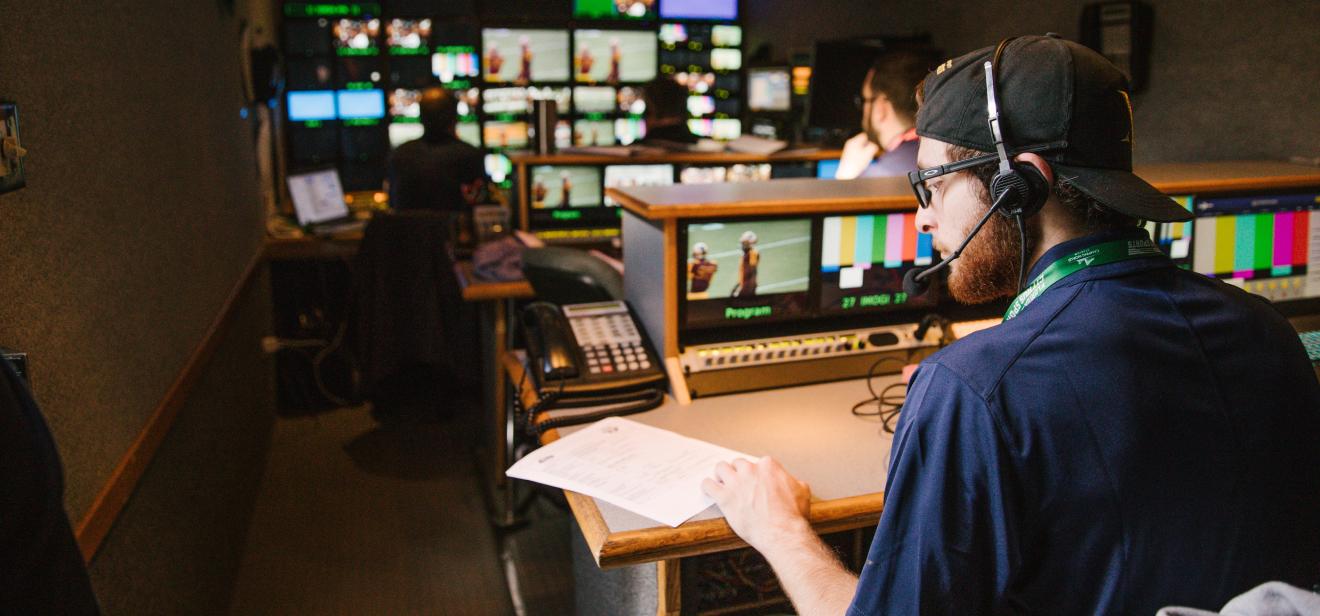 In a control room lit up with TV screens, students wear headsets and monitor shots of a broadcast.