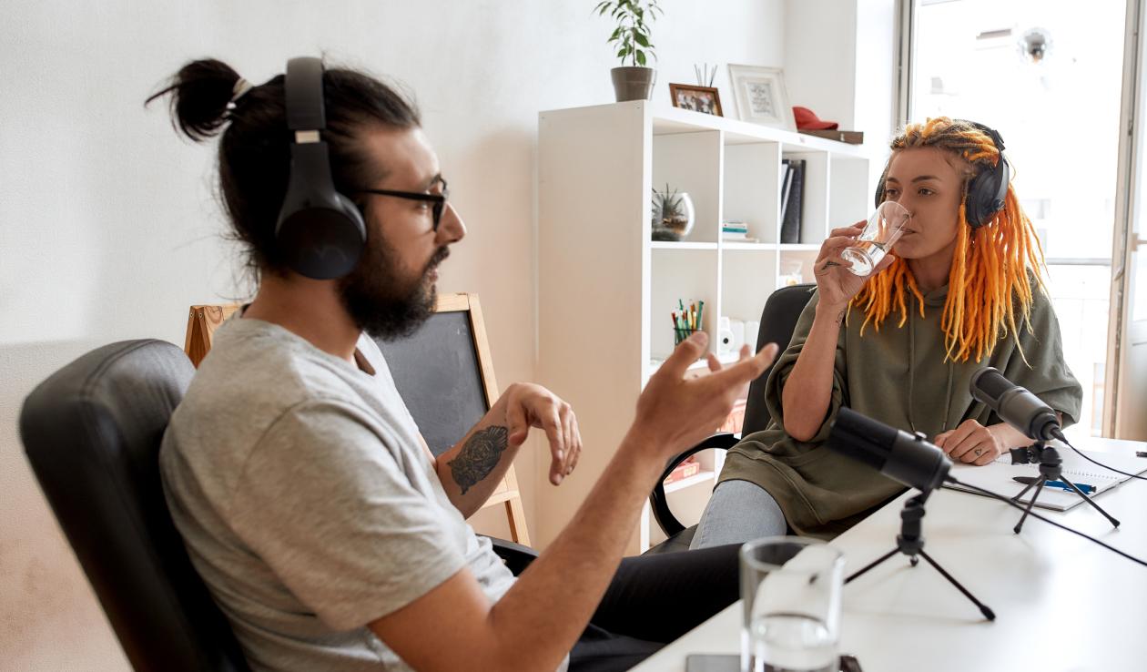 Two students wearing headphones and sitting in front of professional microphones record a podcast.