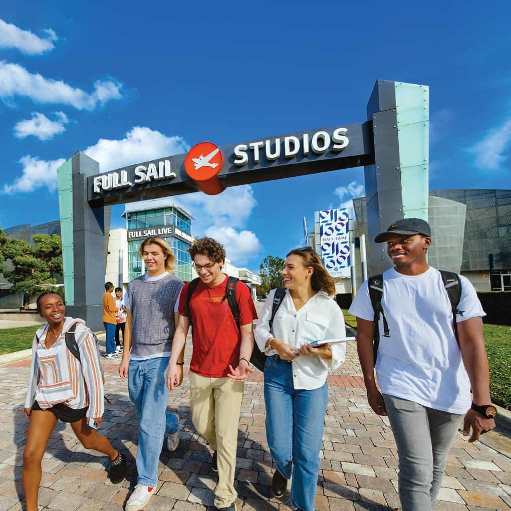 On a bright, sunny campus beneath the Full Sail Studios archway, a group of students with backpacks laugh and walk together.