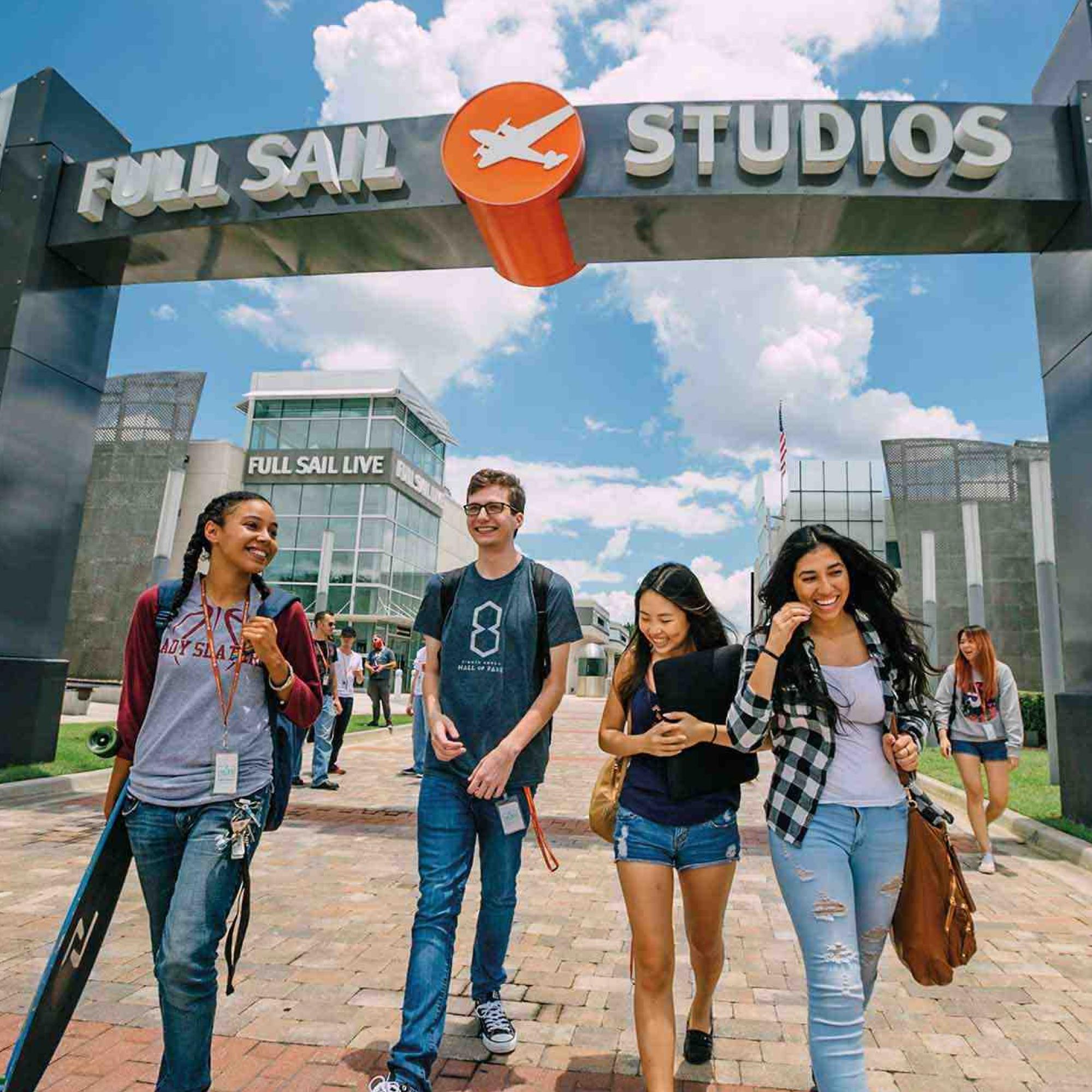 On a bright, sunny campus beneath the Full Sail Studios archway, a group of students with backpacks laugh and walk together.