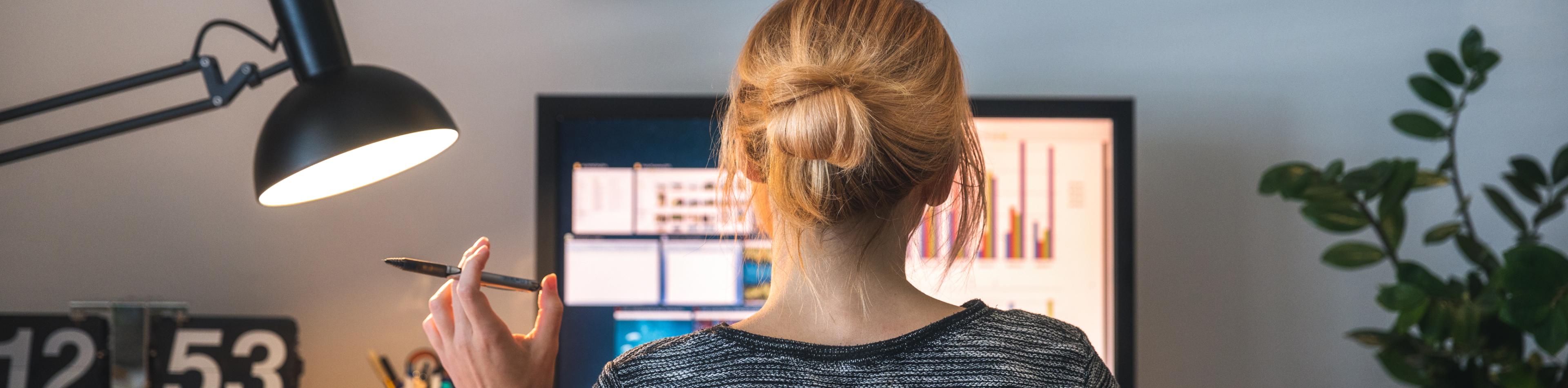 A woman in front of a desktop monitor holds a pen as she contemplates charts and graphs on the screen.