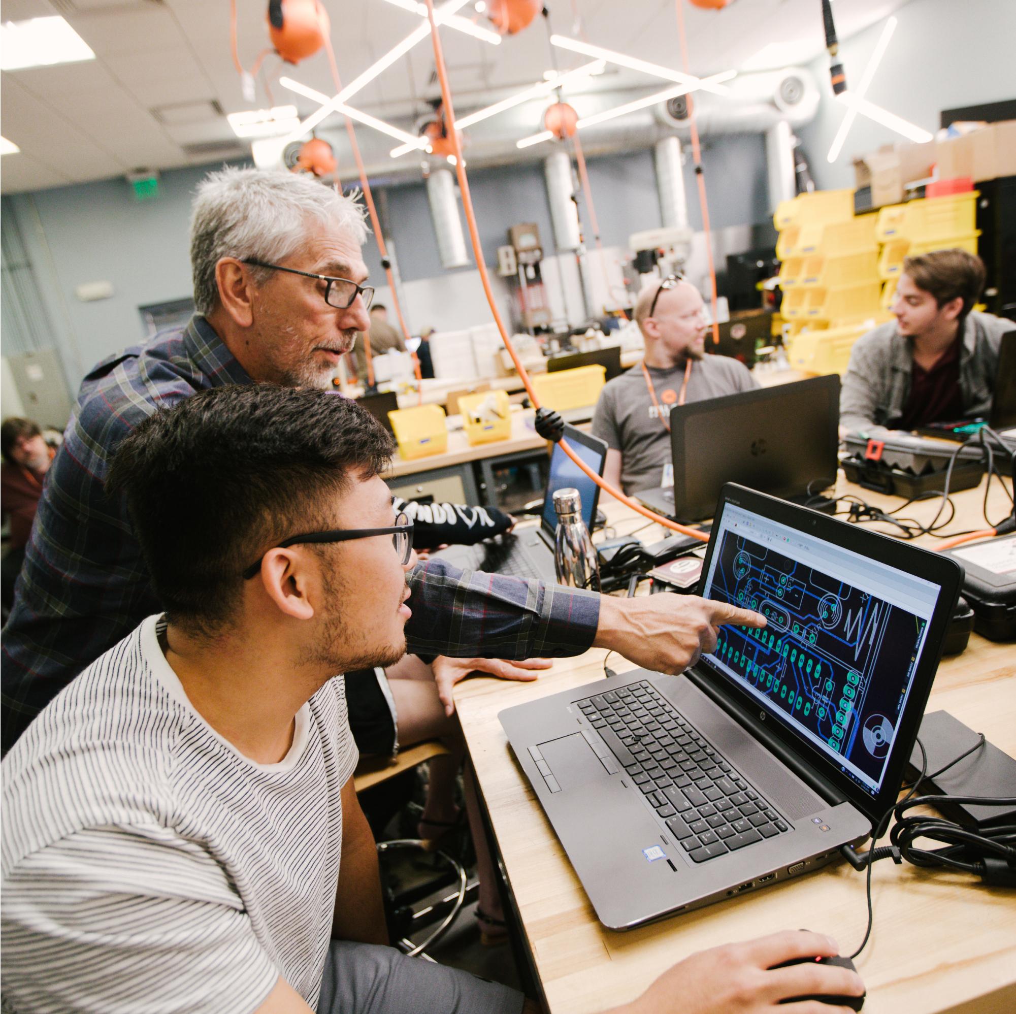 Inside the Fab Lab, an instructor points to a schematic on a screen in front of a student while other students chat in the background.